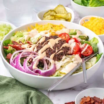 A bowl of salad topped with a cooked burger patty, sliced red onions, cherry tomatoes, shredded cheese, bacon bits, pickles, and creamy dressing. Fork and spoon are placed in the bowl.