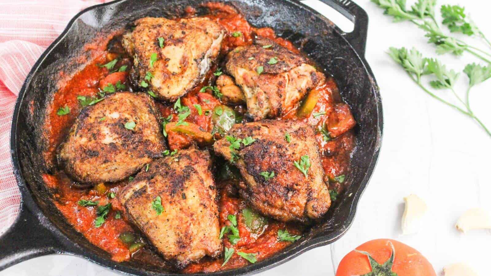 Seasoned chicken thighs cooked in tomato sauce with bell peppers and herbs, served in a cast-iron skillet. Fresh parsley, garlic, and a tomato are nearby.