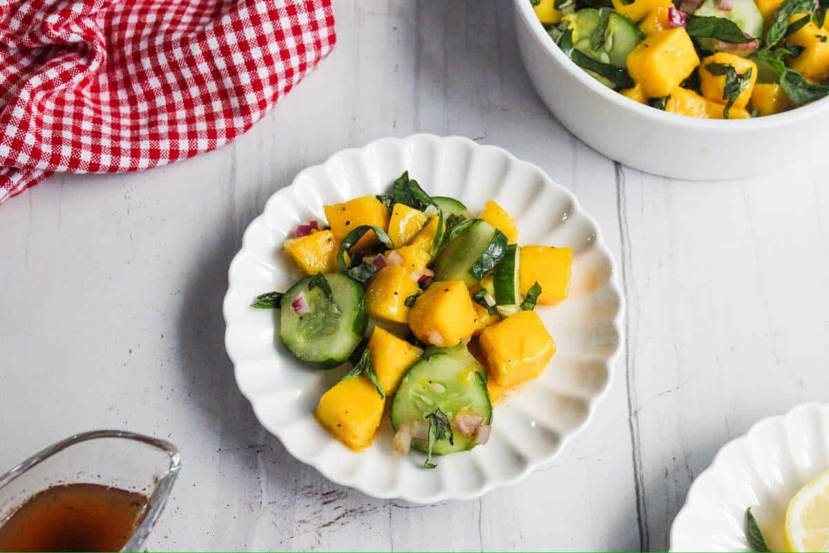 A white plate with a salad made of mango, cucumber, red onion, and basil sits on a light table near a red checkered cloth.