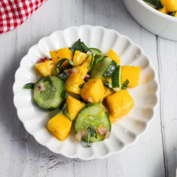 A white scalloped plate holds a salad made of mango chunks, cucumber slices, red onion, and basil on a light wooden surface.