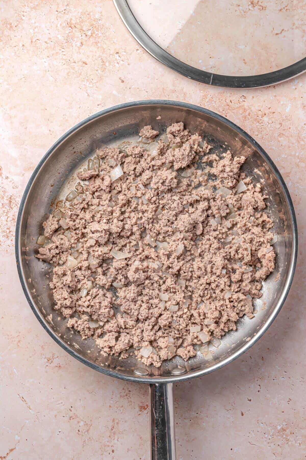Ground meat and chopped onions cooking in a stainless steel frying pan on a light-colored countertop.