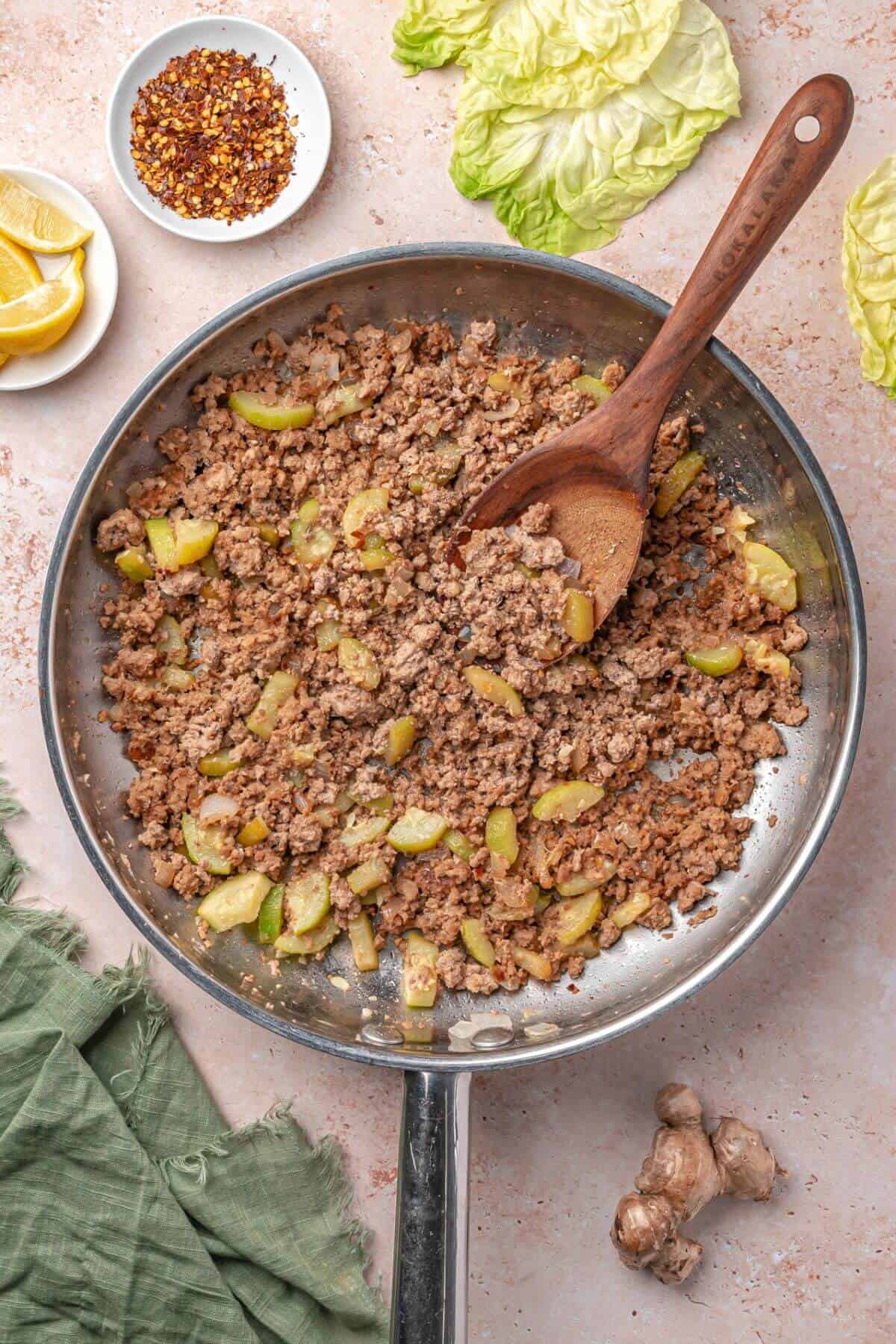 Ground meat and chopped vegetables cooked in a skillet with a wooden spoon, surrounded by lettuce leaves, lemon wedges, red pepper flakes, and a green cloth.