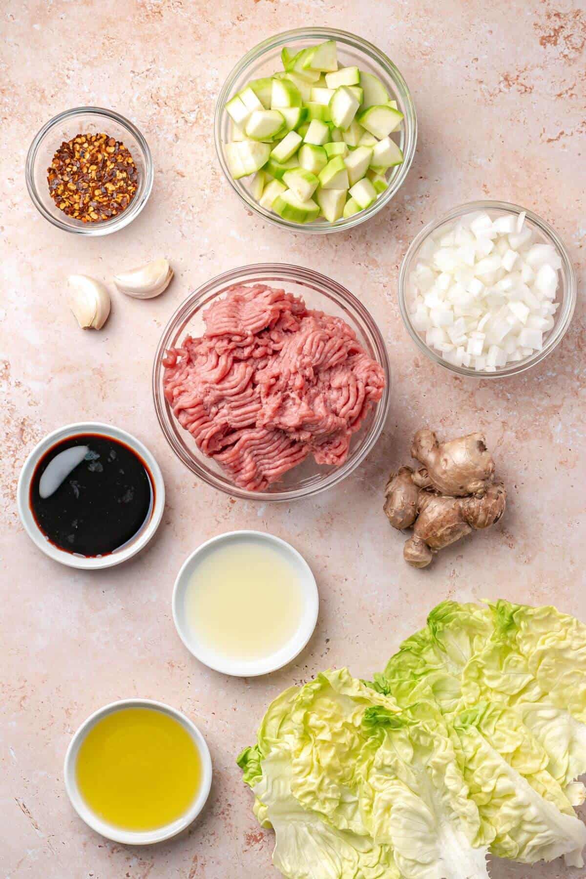 Bowls of ground meat, diced zucchini, chopped onion, soy sauce, lemon juice, olive oil, red pepper flakes, garlic, ginger, and lettuce leaves arranged on a light surface.