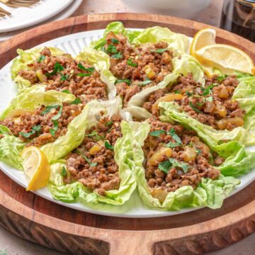 A plate of lettuce wraps filled with seasoned ground meat and garnished with chopped herbs, served with lemon wedges.