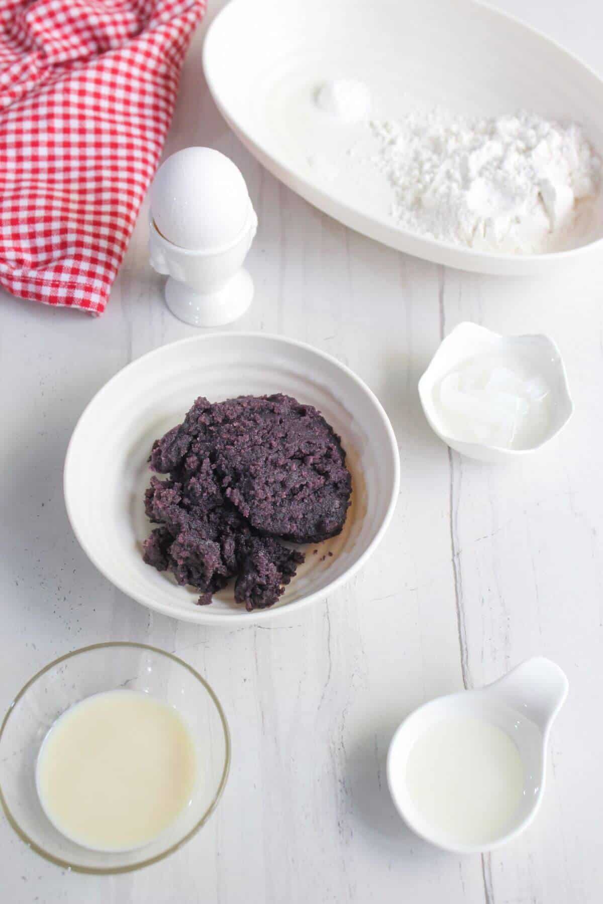 Ingredients for baking displayed on a white surface include flour, an egg, a red bean paste, a red checkered cloth, and small bowls of liquid and solid ingredients.