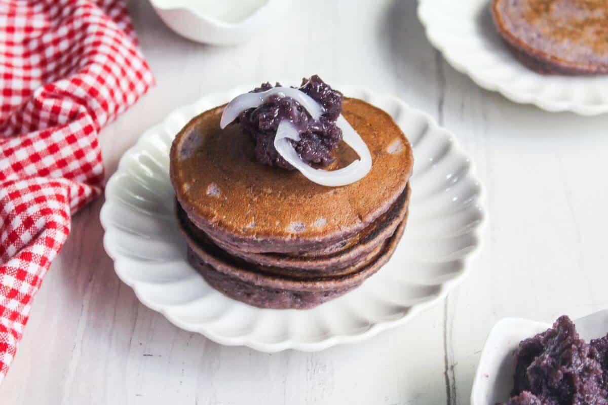 A stack of purple pancakes topped with a dark spread and white coconut strips on a white plate, with a red checkered cloth nearby.