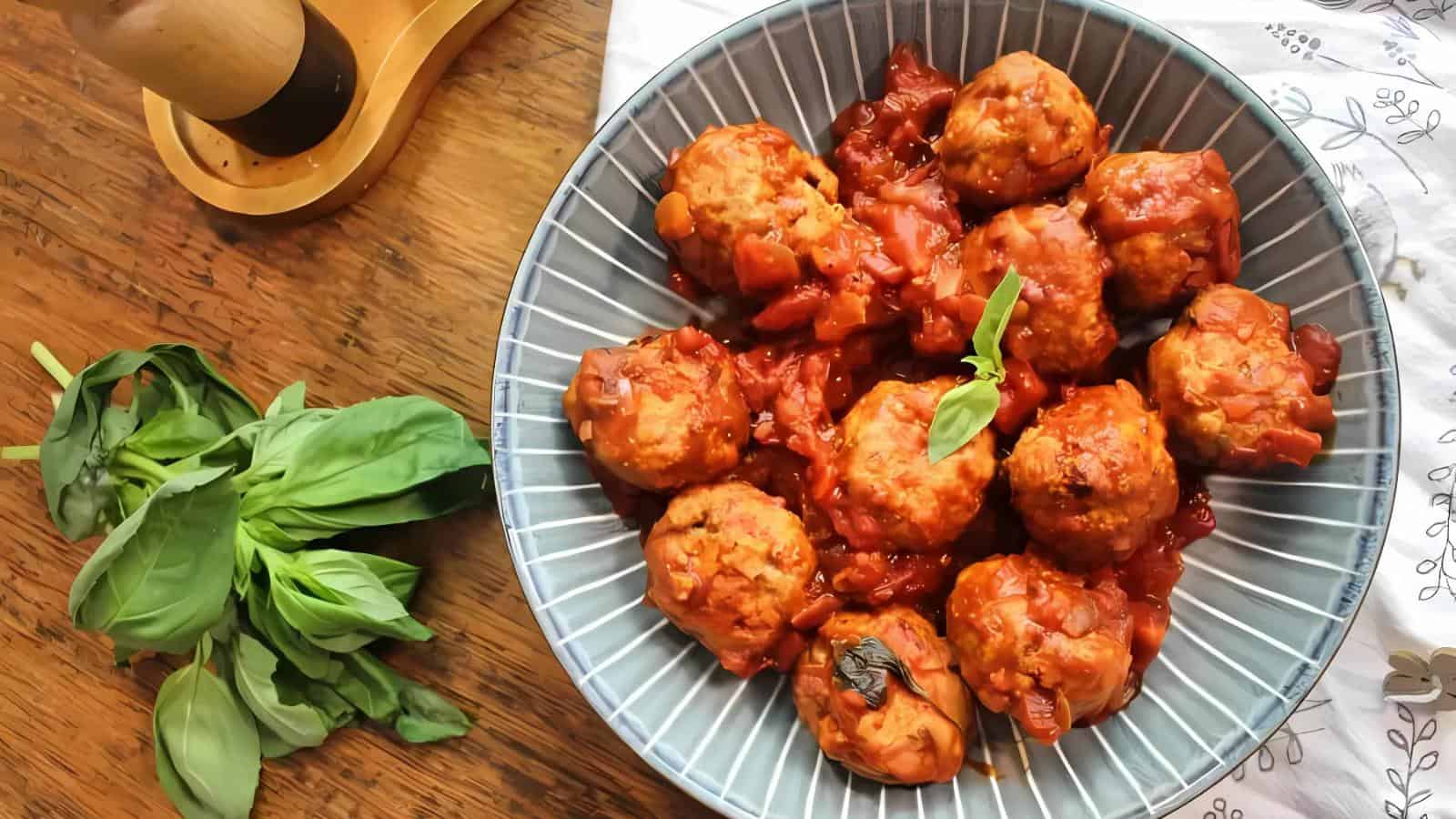 A blue bowl filled with meatballs in tomato sauce is on a wooden table next to a bunch of fresh basil and a pepper grinder.