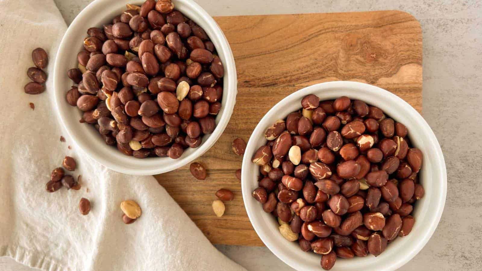 Two white bowls filled with shelled peanuts are placed on a wooden cutting board, with a few loose peanuts on a white cloth nearby.