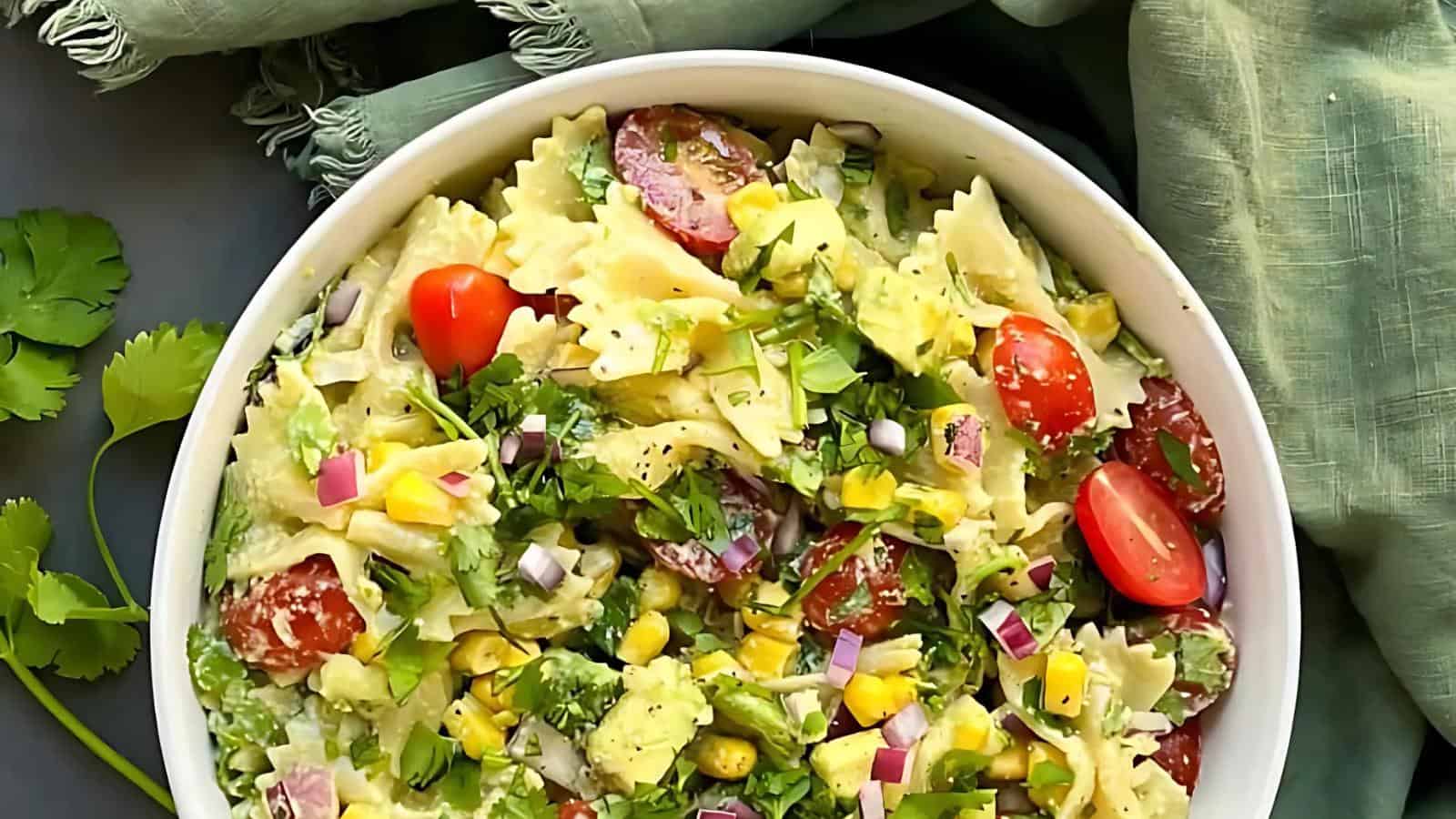 A bowl of pasta salad with bowtie pasta, cherry tomatoes, corn, red onion, avocado, and chopped cilantro, set on a green cloth.