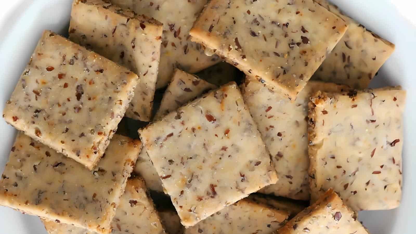 A close-up of square, light brown crackers with visible seeds and grains, arranged in a white bowl.