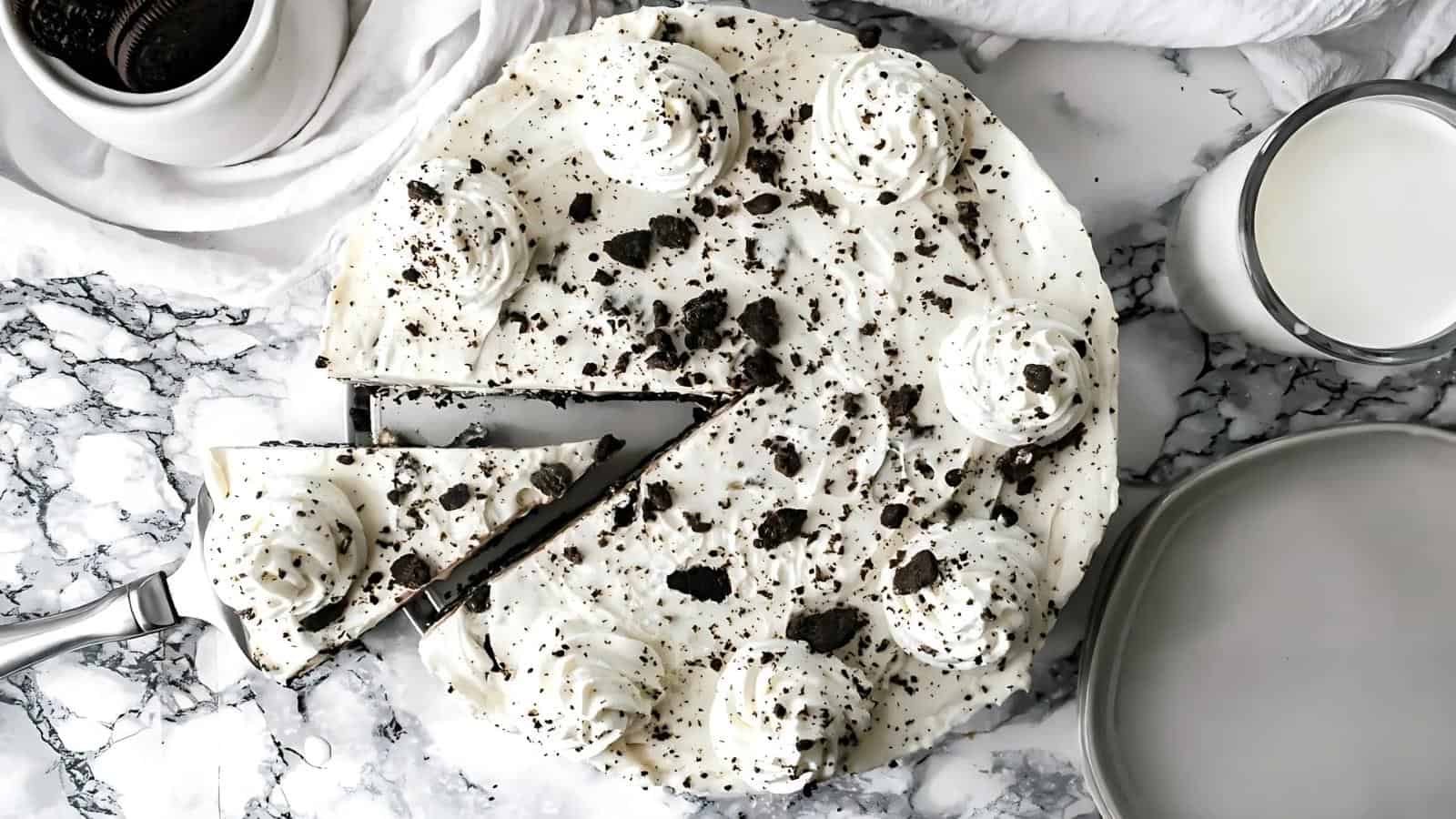 A cookies and cream cheesecake with whipped cream swirls and cookie crumbs on top, partially sliced, on a marble surface next to a glass of milk and a plate.