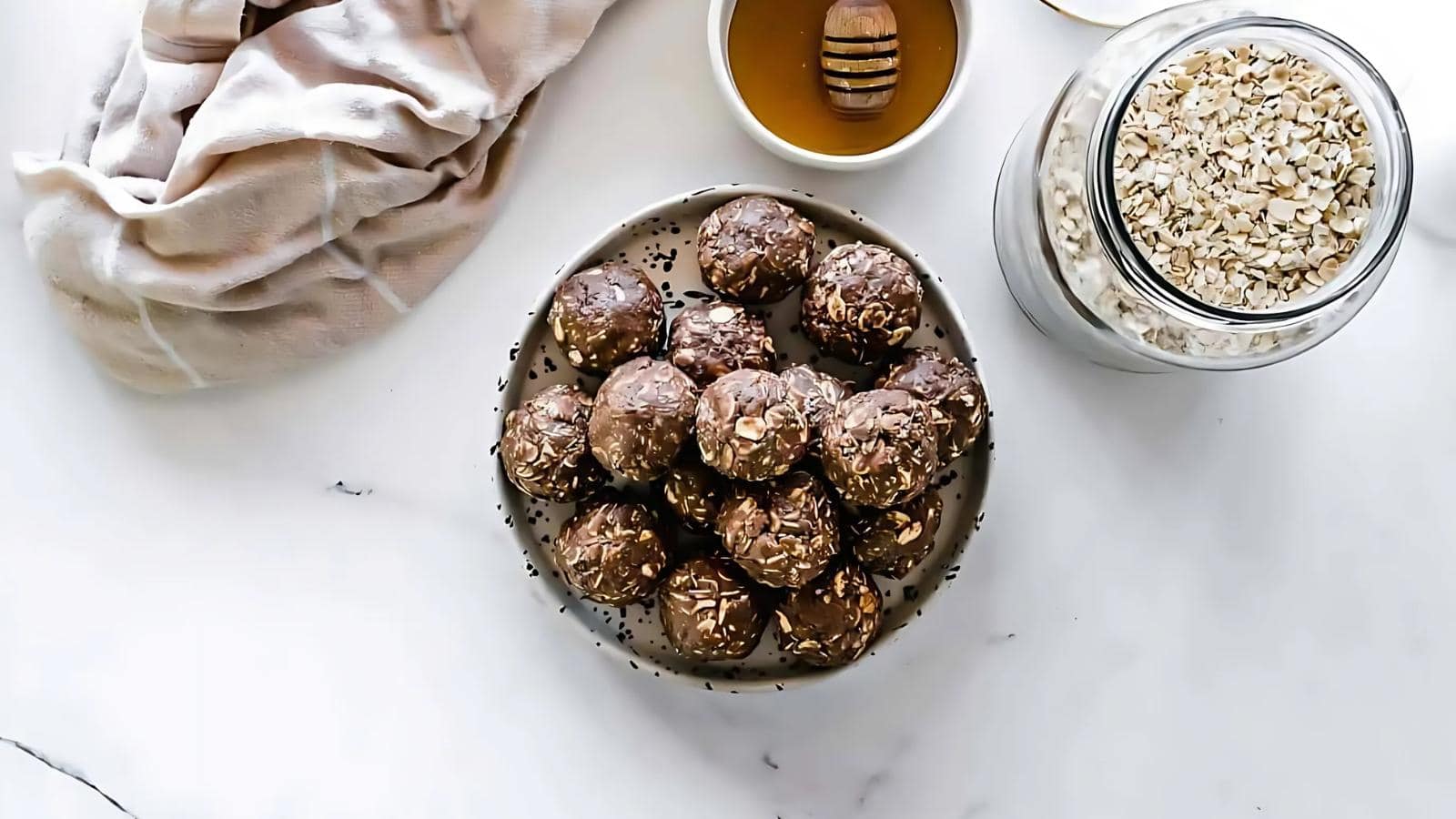 A plate of chocolate oat energy balls sits on a white surface next to a jar of oats, a bowl of honey with a honey dipper, and a beige cloth.