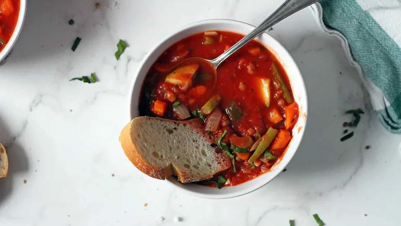 A bowl of vegetable soup with a slice of bread and a spoon, placed on a white marble surface with scattered herbs.