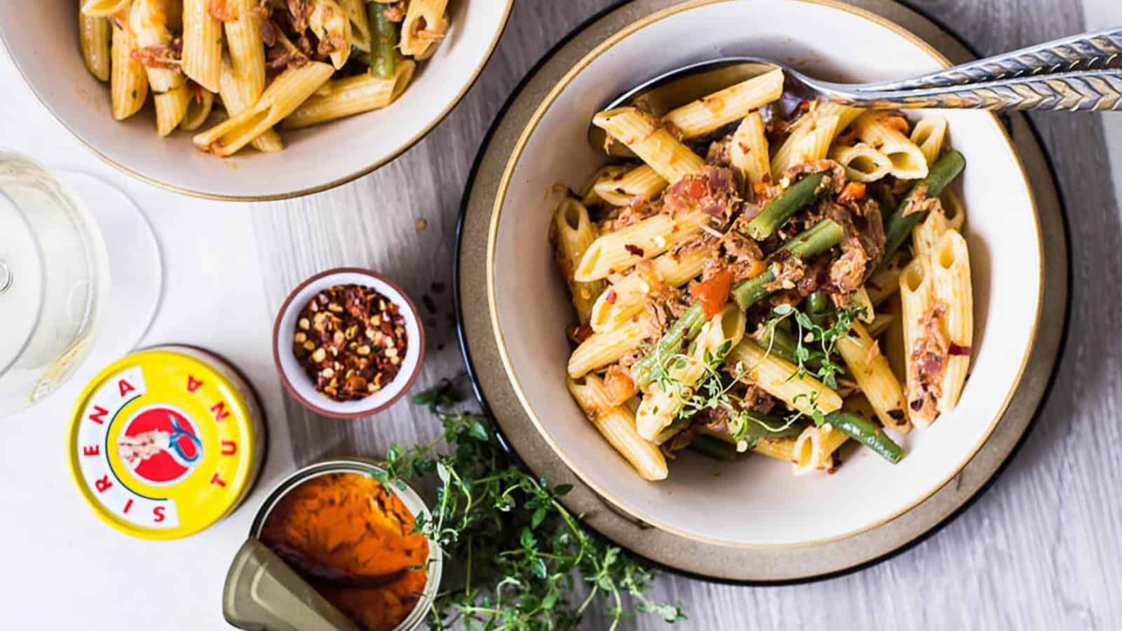 A bowl of penne pasta with green beans and tomato sauce, garnished with fresh herbs, sits beside a can of tuna, red pepper flakes, and another bowl of pasta on a wooden table.