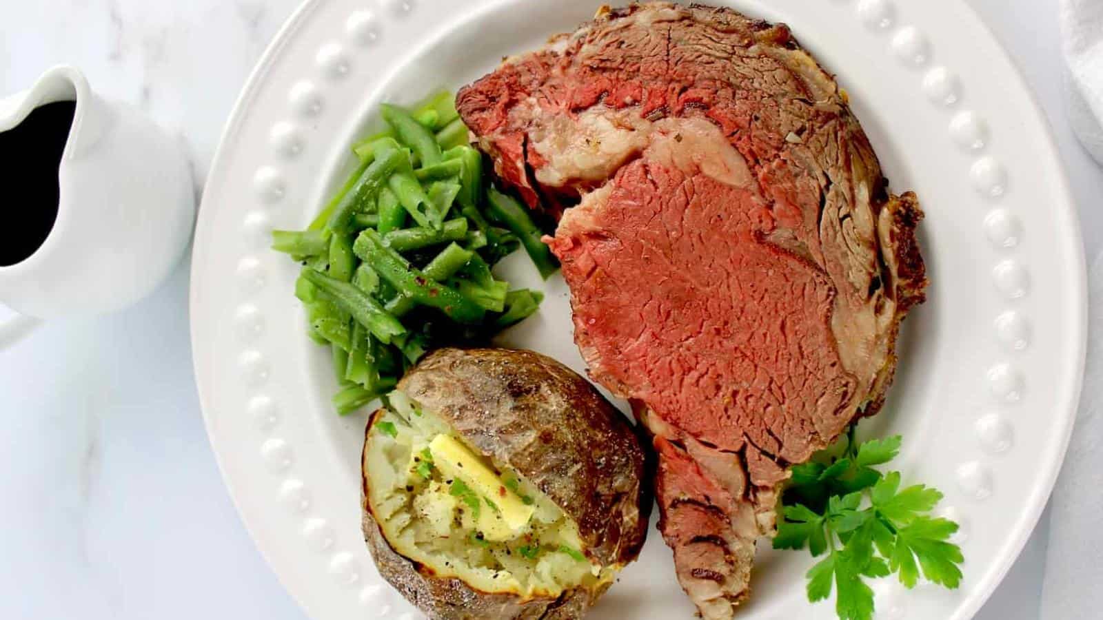 A plate with a slice of rare roast beef, green beans, and a baked potato with butter, garnished with parsley. A small pitcher of sauce is next to the plate.