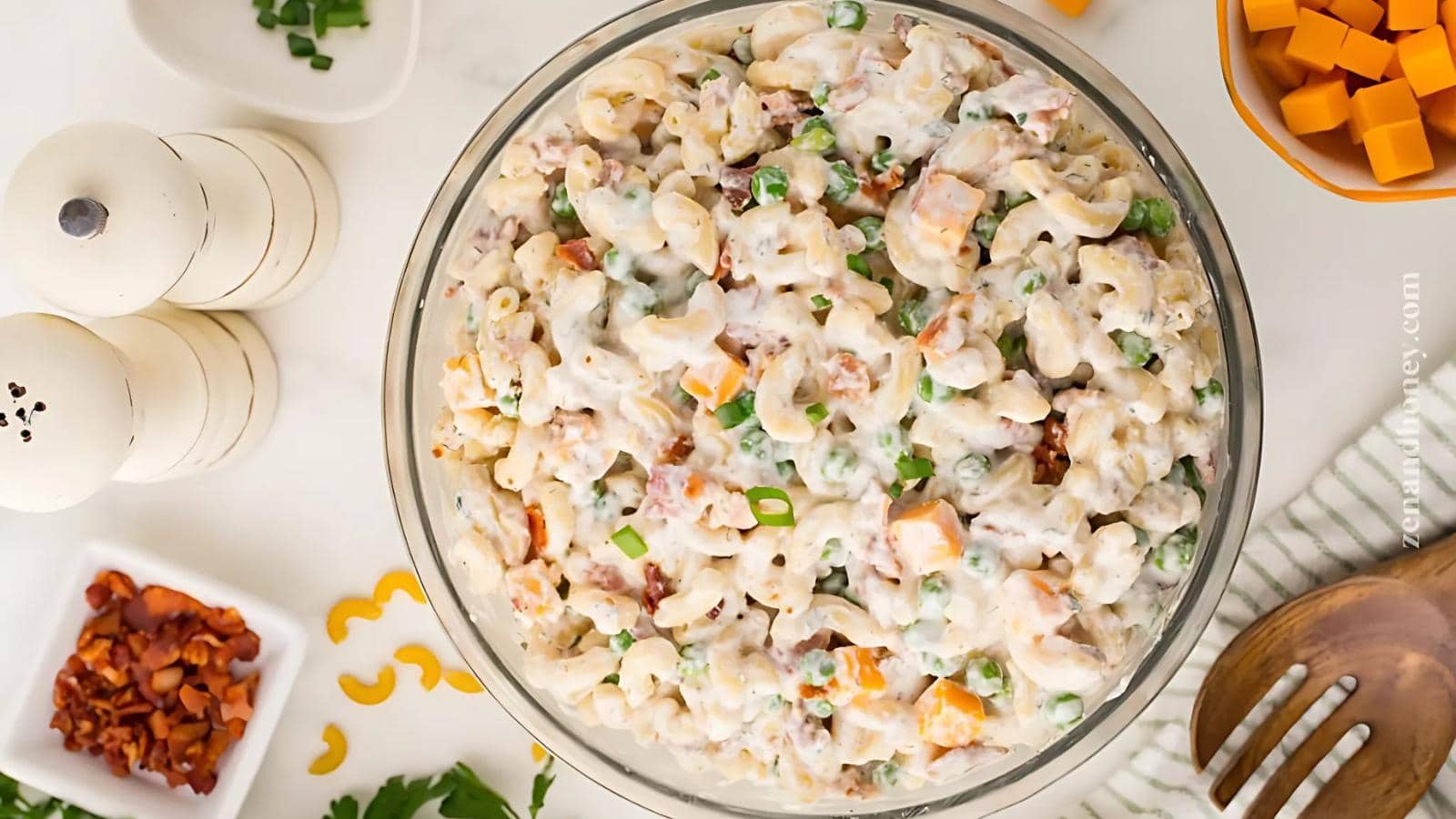 A glass bowl filled with macaroni salad mixed with peas, bacon, green onions, and cheese cubes sits on a white surface, surrounded by ingredients and utensils.