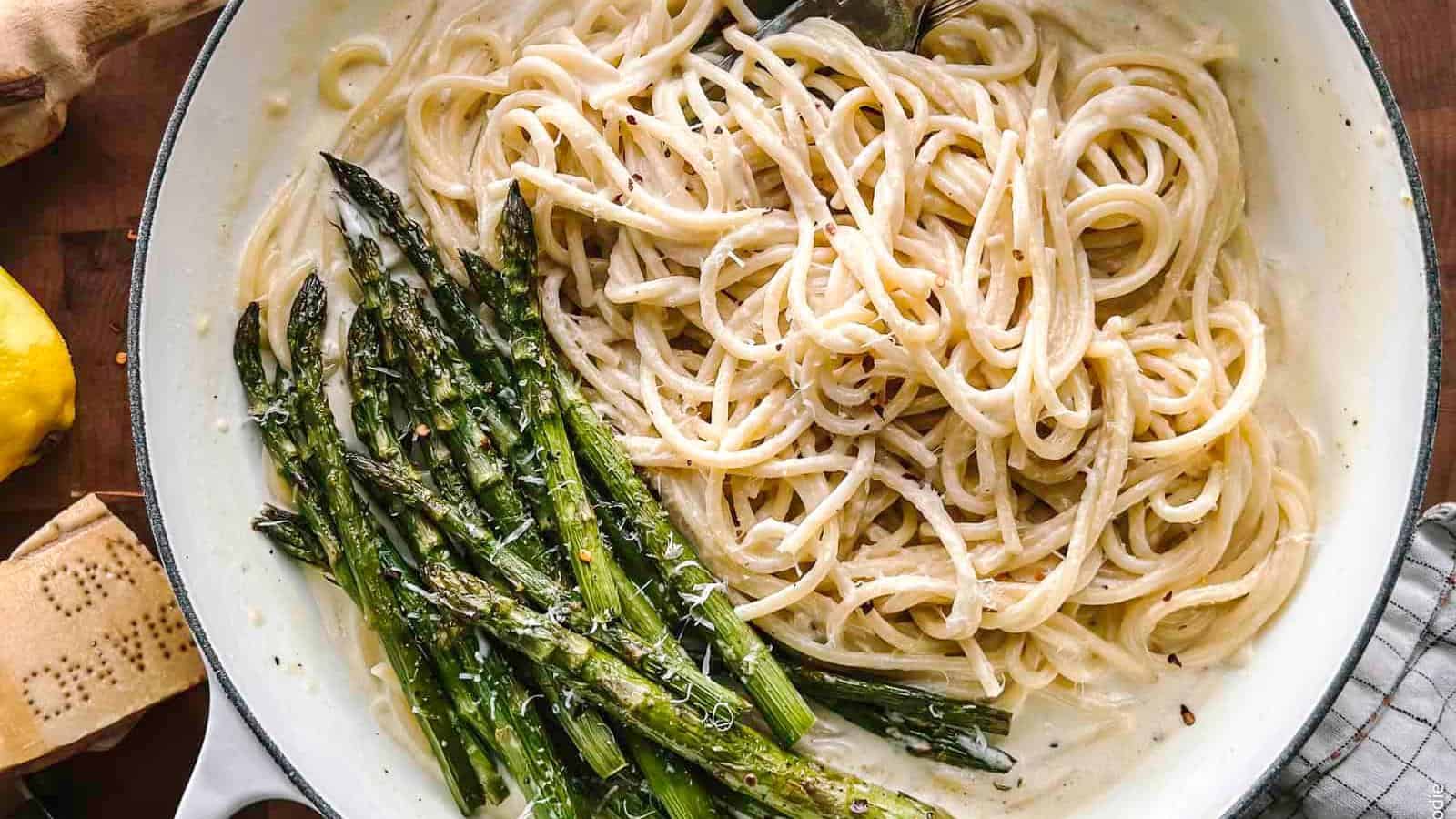 Cooked spaghetti in a creamy white sauce with roasted asparagus on the side, served in a large white pan.