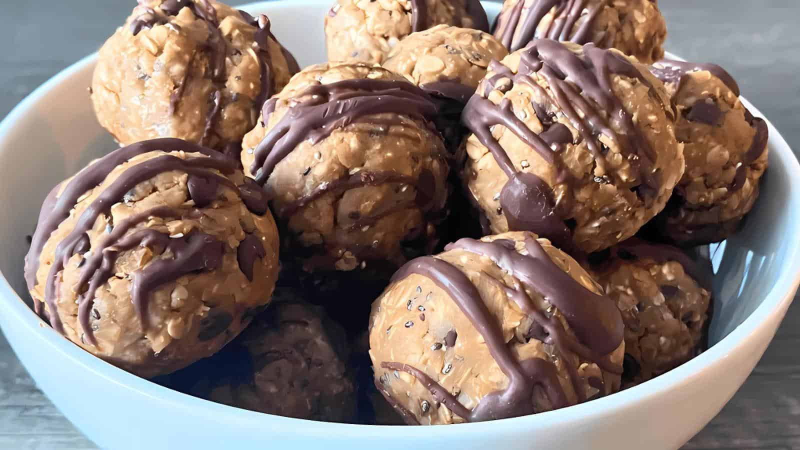 A white bowl filled with round peanut butter oat energy balls drizzled with chocolate, on a gray wooden surface.