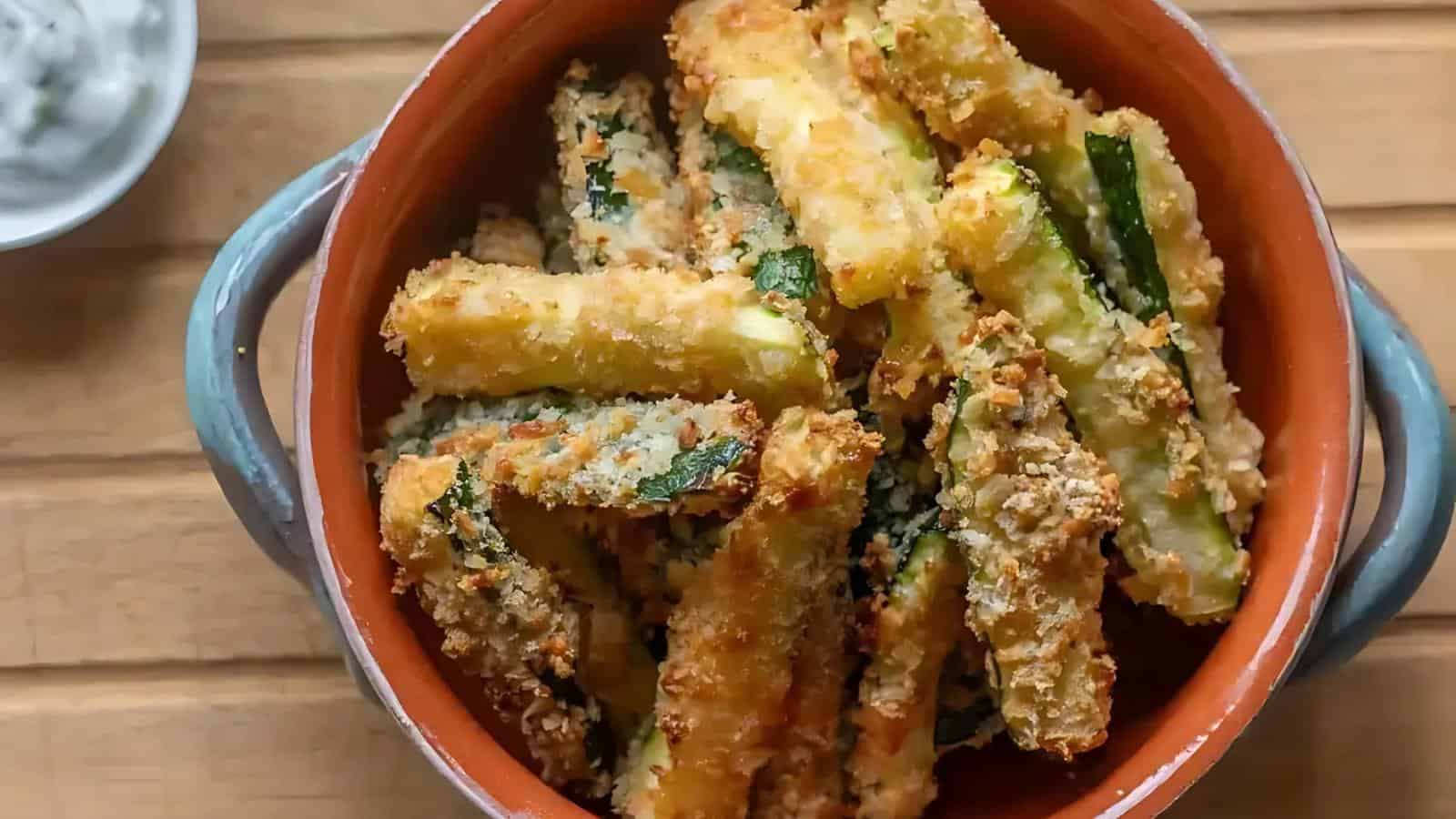 A ceramic bowl filled with crispy, breaded zucchini fries sits on a wooden surface.