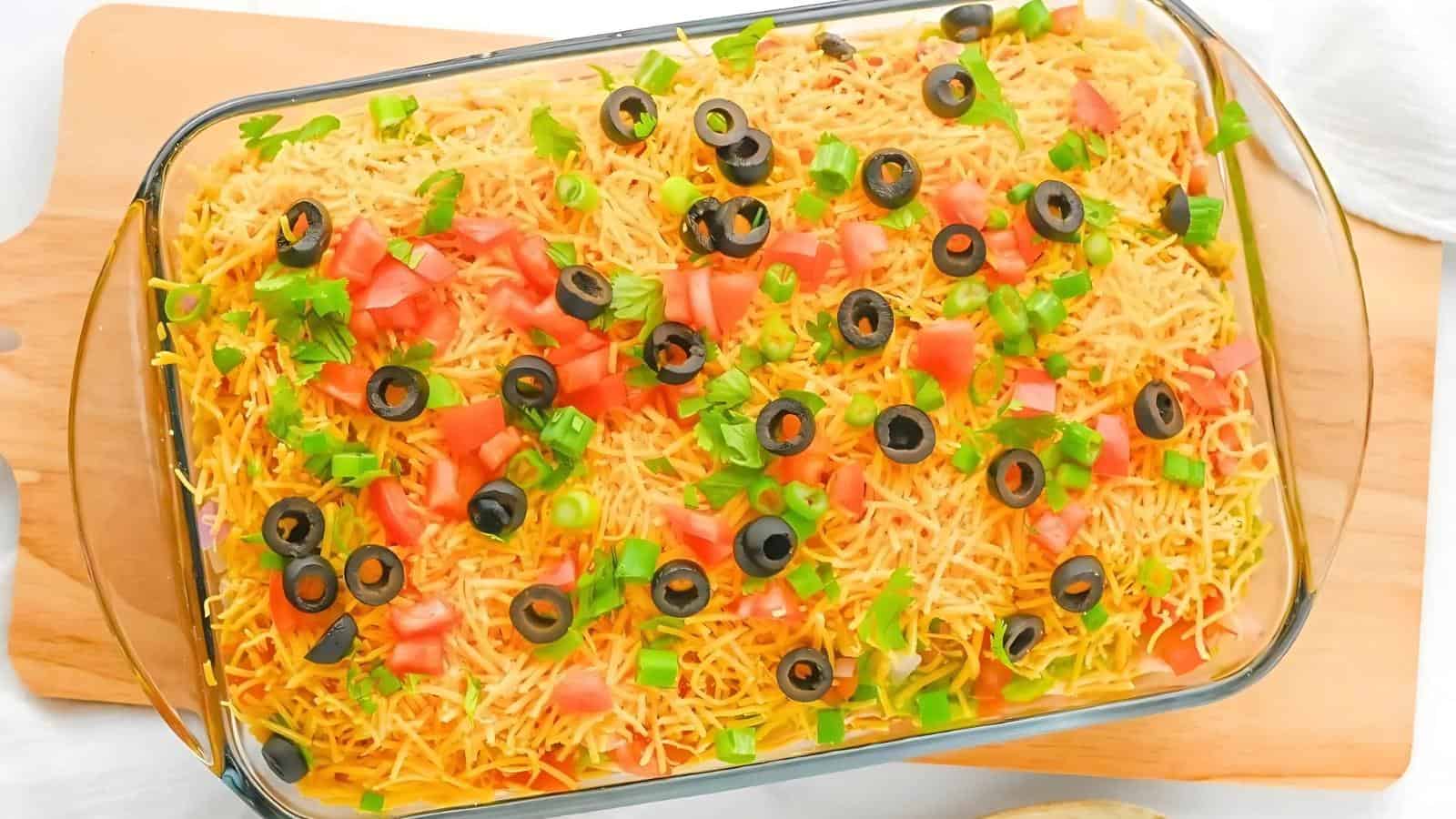 A glass baking dish filled with a layered dip topped with shredded cheese, diced tomatoes, sliced black olives, and chopped green onions on a wooden cutting board.