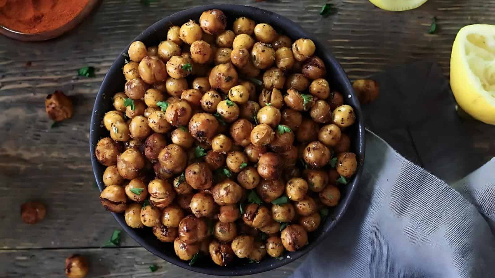 A bowl of roasted chickpeas garnished with herbs sits on a wooden table next to a gray napkin and a lemon half.