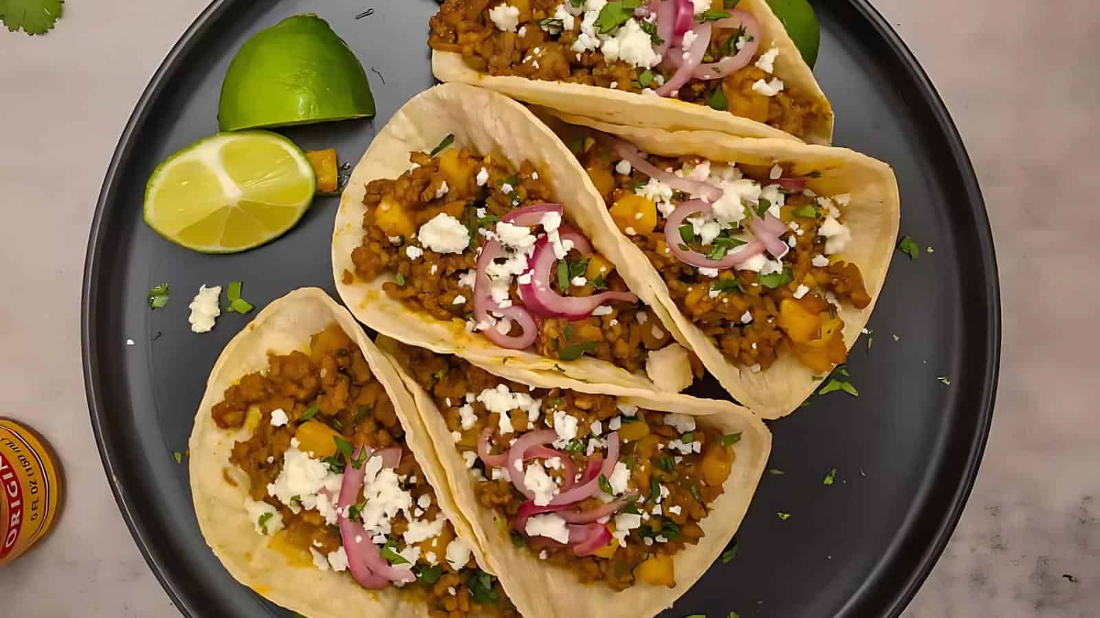 Four tacos filled with spiced ground filling, diced vegetables, pickled onions, and crumbled cheese are arranged on a black plate with lime wedges on the side.