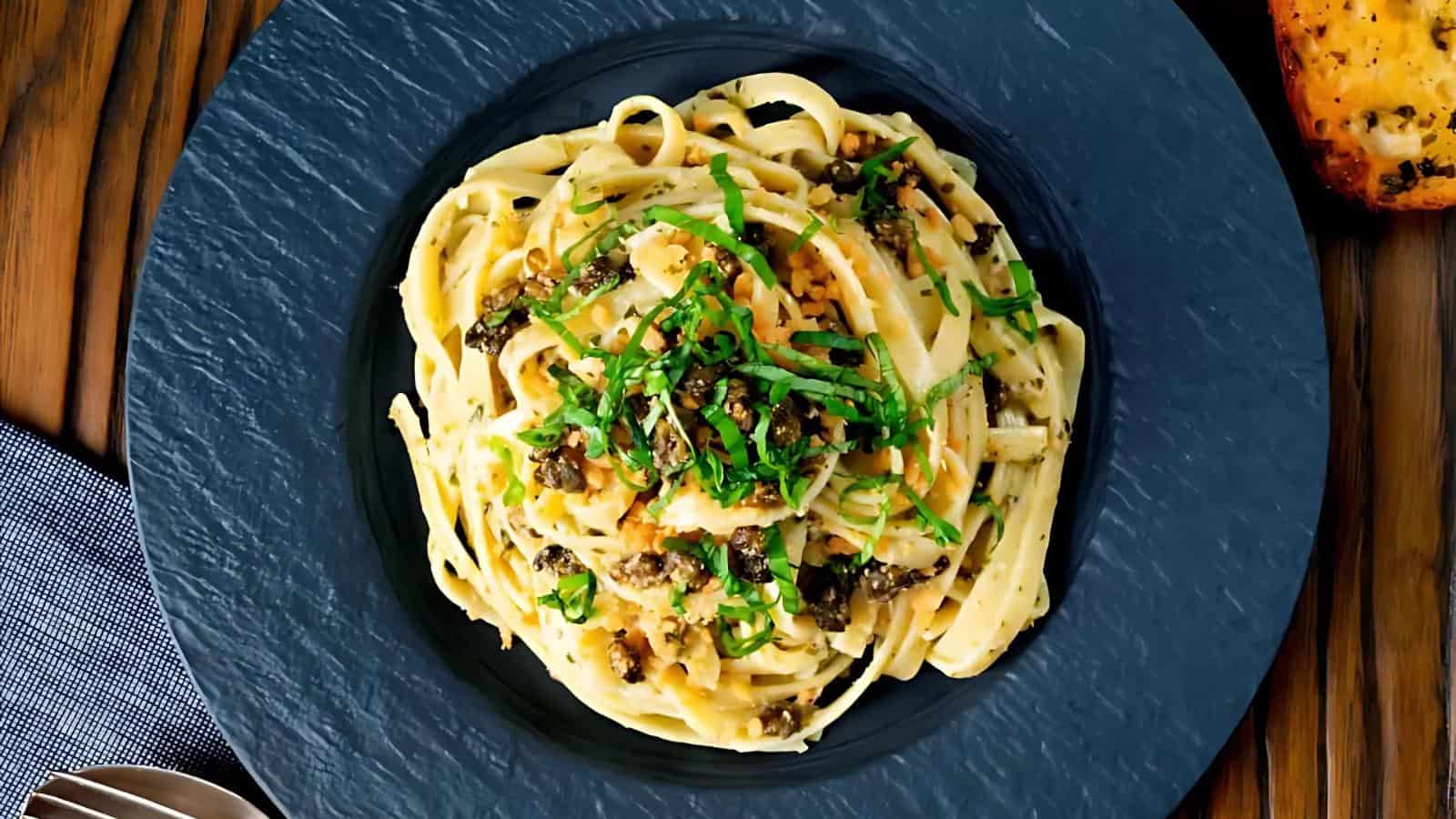 A plate of fettuccine pasta topped with chopped herbs and a savory sauce, served on a dark round plate.