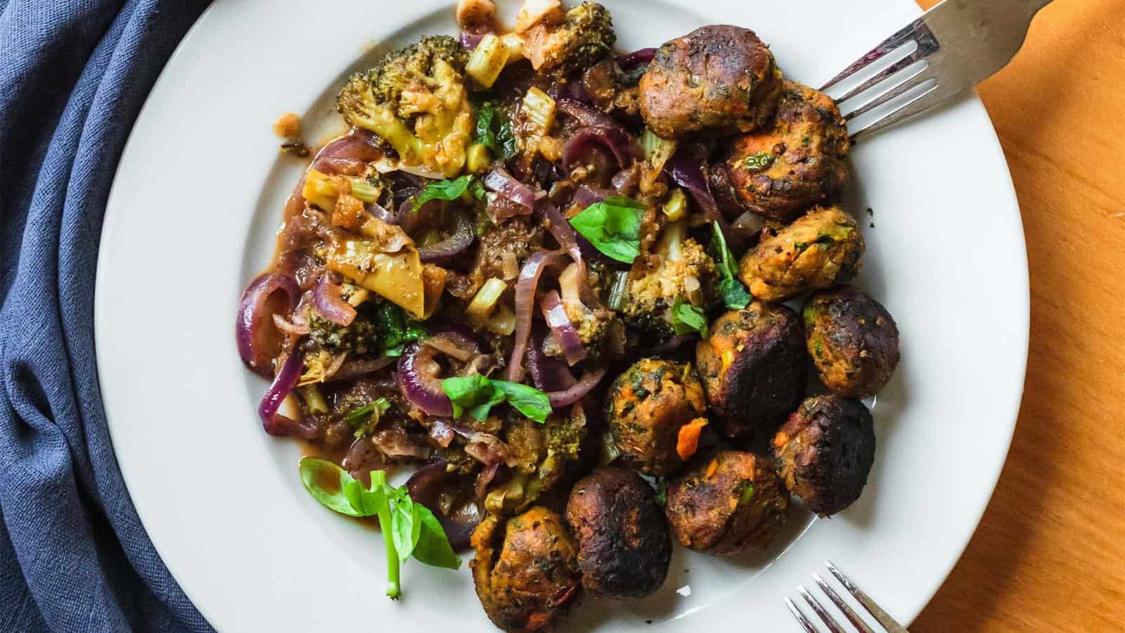 A white plate with vegetable stew on one side and a serving of browned vegetable balls on the other, garnished with fresh basil leaves. A fork and knife sit at the edge of the plate.