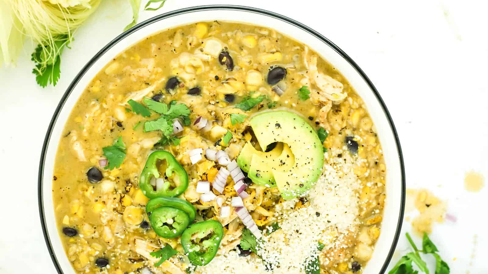 A bowl of chicken chili topped with avocado slices, jalapeños, chopped red onion, cilantro, and grated cheese.