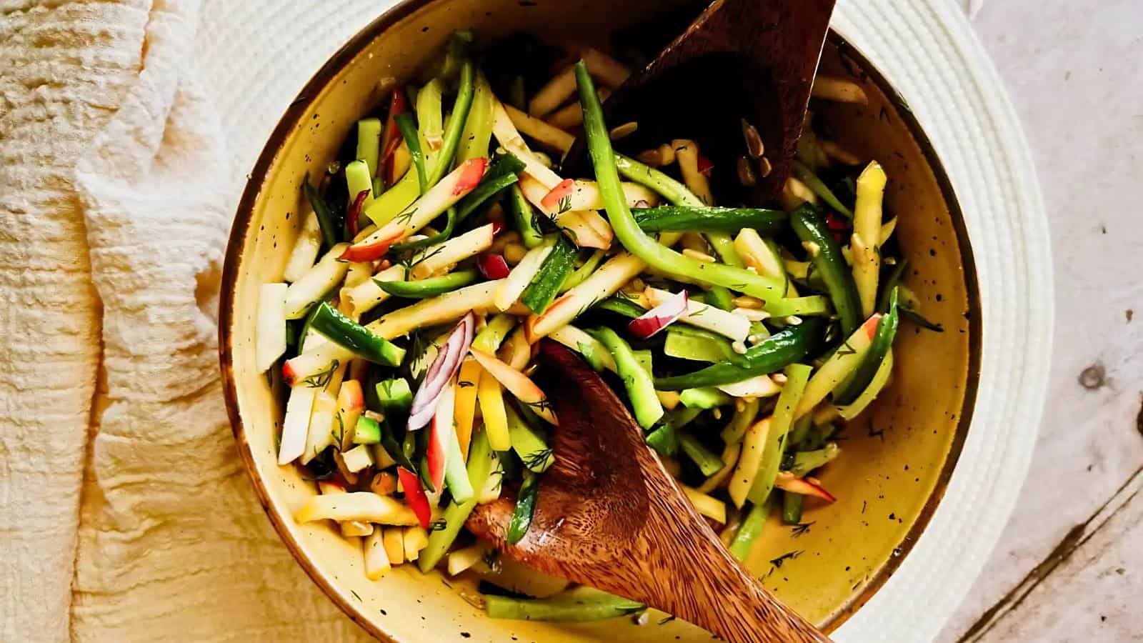A bowl of julienned vegetables, including cucumbers, apples, and onions, tossed together with herbs, with two wooden salad servers.