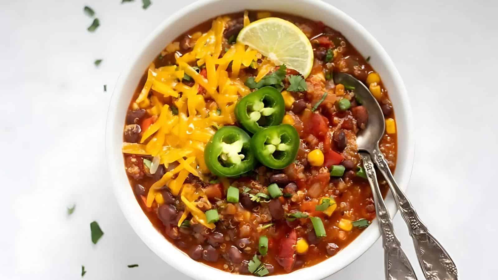 A bowl of chili topped with shredded cheddar cheese, sliced jalapeños, chopped herbs, and a lime wedge, with two spoons placed inside.