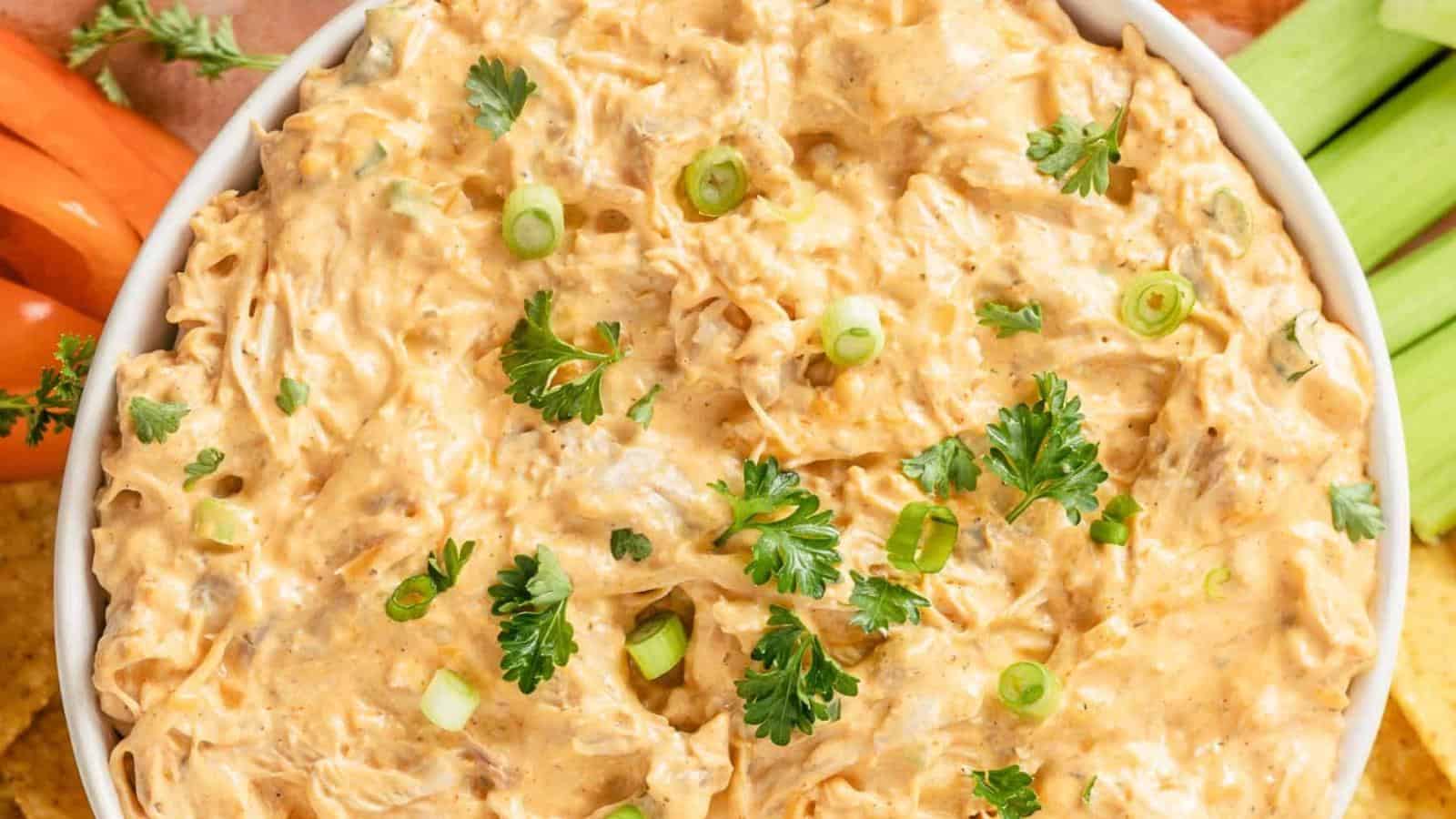 A bowl of creamy buffalo chicken dip garnished with chopped green onions and parsley, surrounded by celery sticks and orange vegetables.