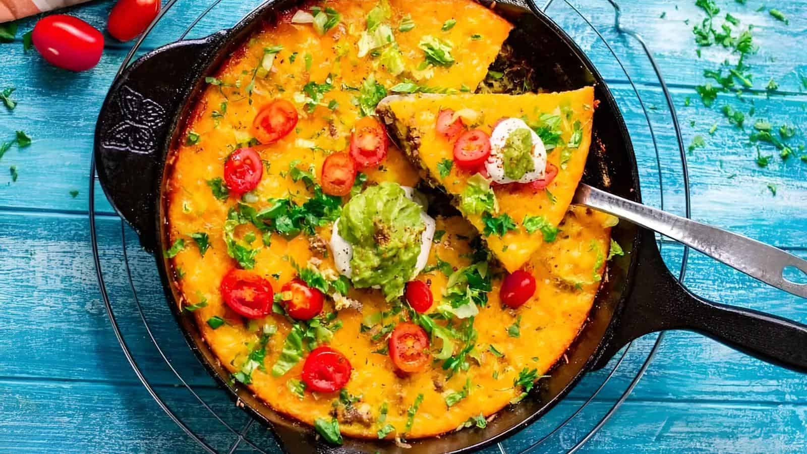 A cast iron skillet filled with a baked cheesy casserole topped with cherry tomatoes, cilantro, guacamole, and sour cream, with one slice being lifted out.