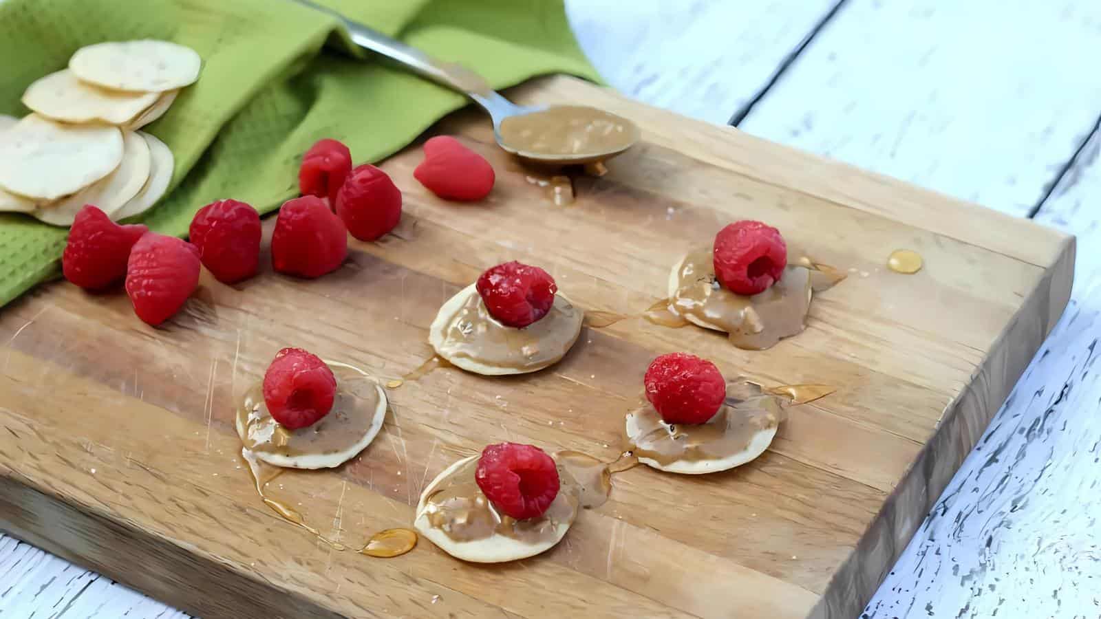A wooden board with mini rice cakes topped with peanut butter, raspberries, and drizzled honey; extra raspberries, rice cakes, and a spoon nearby.