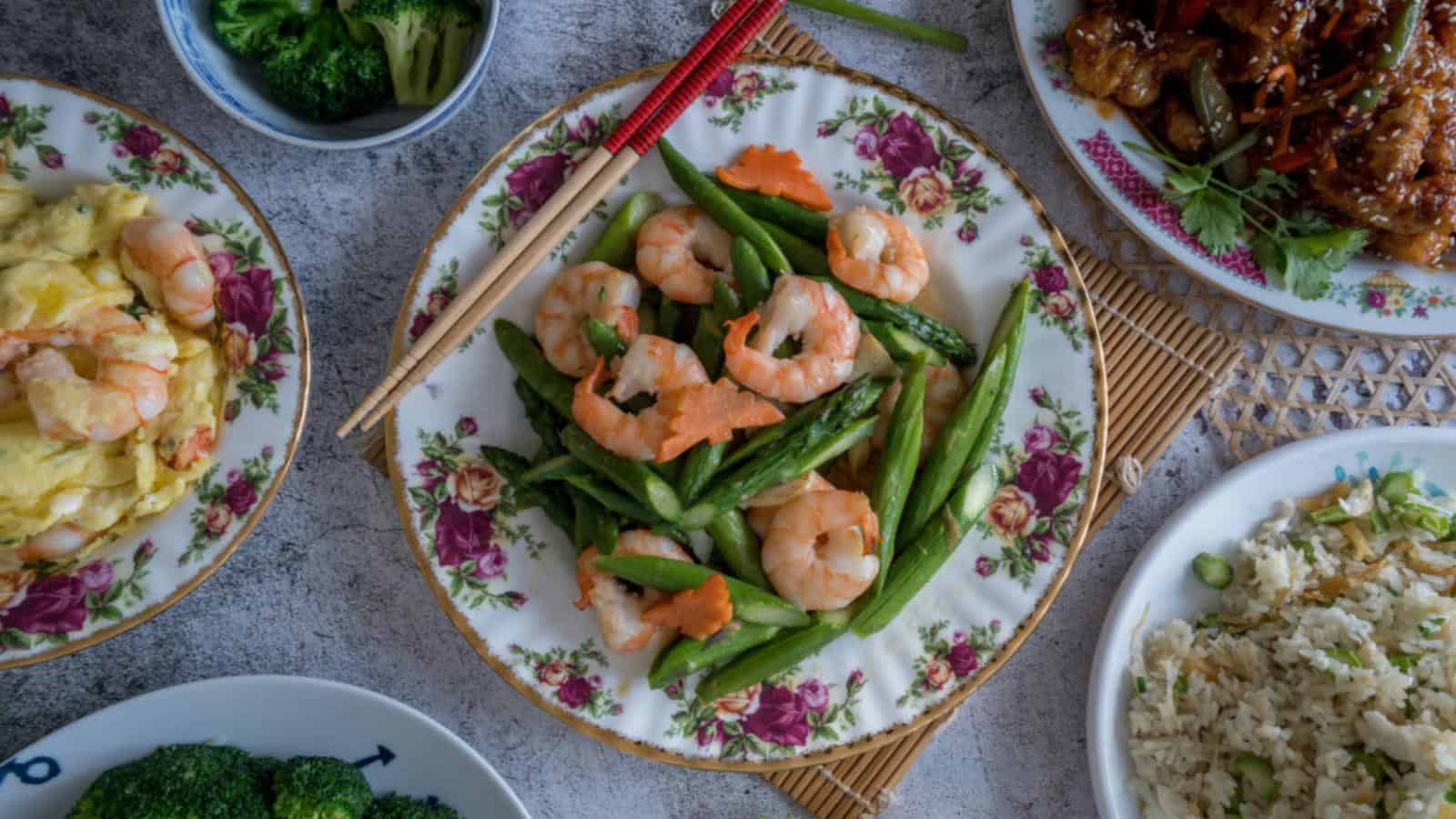 A plate of shrimp and green beans, surrounded by bowls of broccoli, stir-fried rice, an egg dish with shrimp, and a meat dish, all served on floral-patterned dishes.