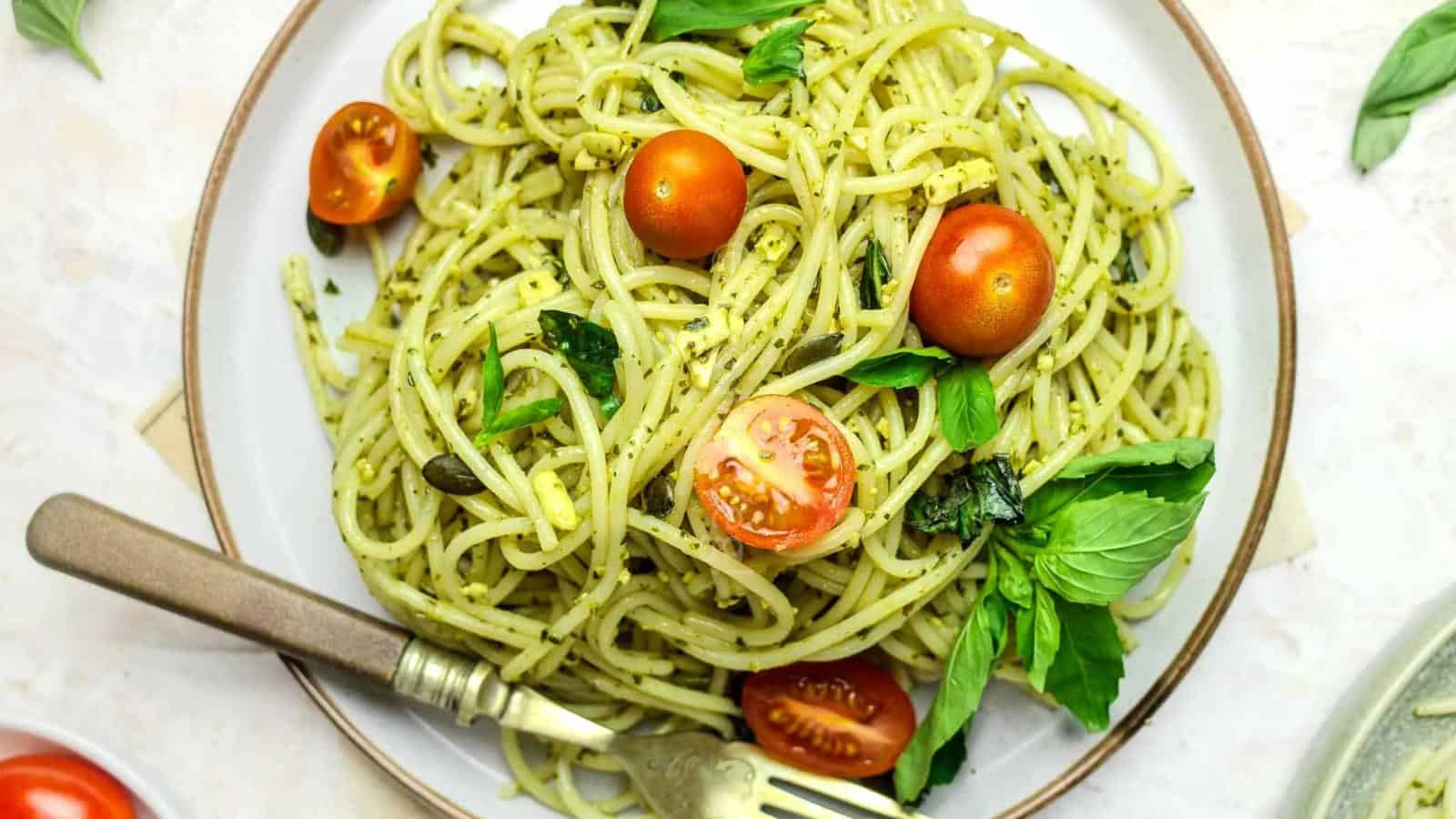 A plate of spaghetti with green pesto sauce, garnished with fresh basil leaves and halved cherry tomatoes, with a fork resting on the side.