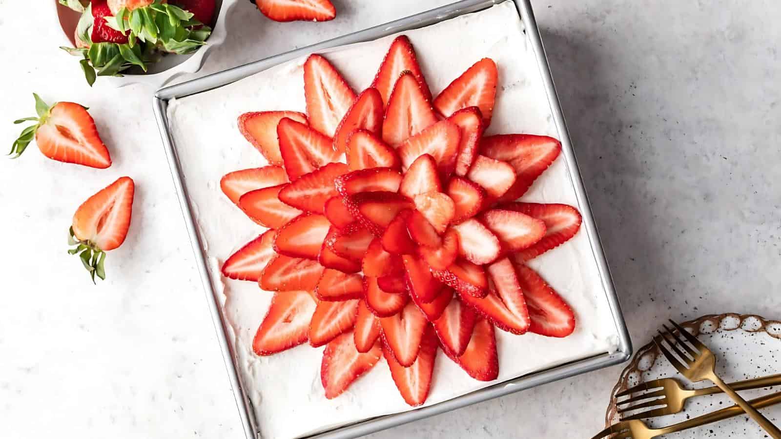 A square cake with white frosting is topped with neatly arranged sliced strawberries in a flower pattern. A bowl of strawberries and gold utensils are nearby.