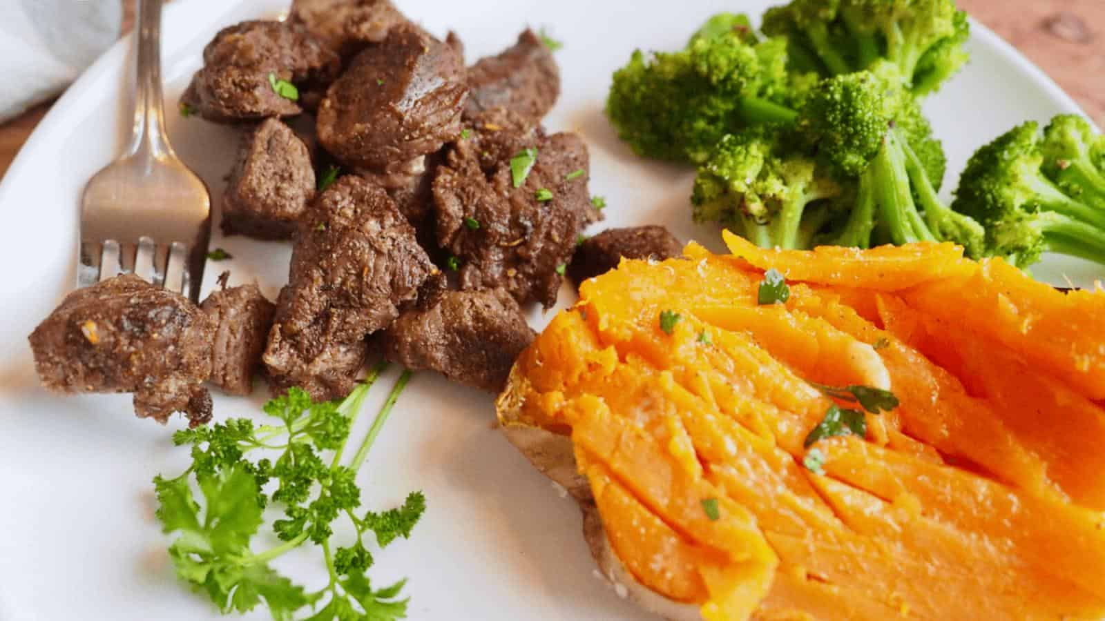A white plate with cooked beef cubes, steamed broccoli, a baked sweet potato, and a sprig of parsley, with a fork resting on the plate.