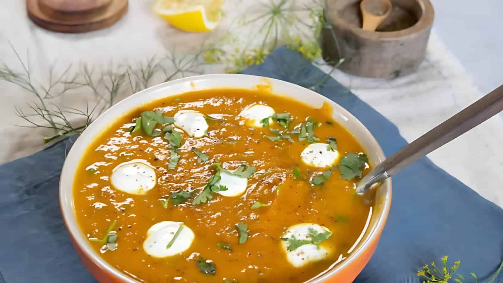 Bowl of orange soup garnished with dollops of cream and chopped herbs, with a spoon inside. In the background are a lemon wedge, a wooden container, and fresh herbs.