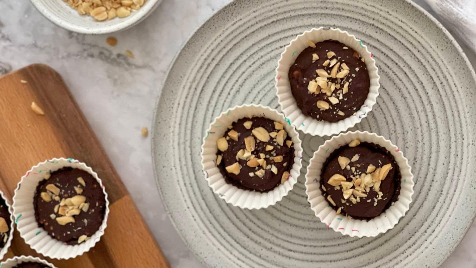 Four chocolate cups topped with chopped peanuts are arranged on a grey plate, with another cup and a bowl of peanuts visible nearby on a marble surface.