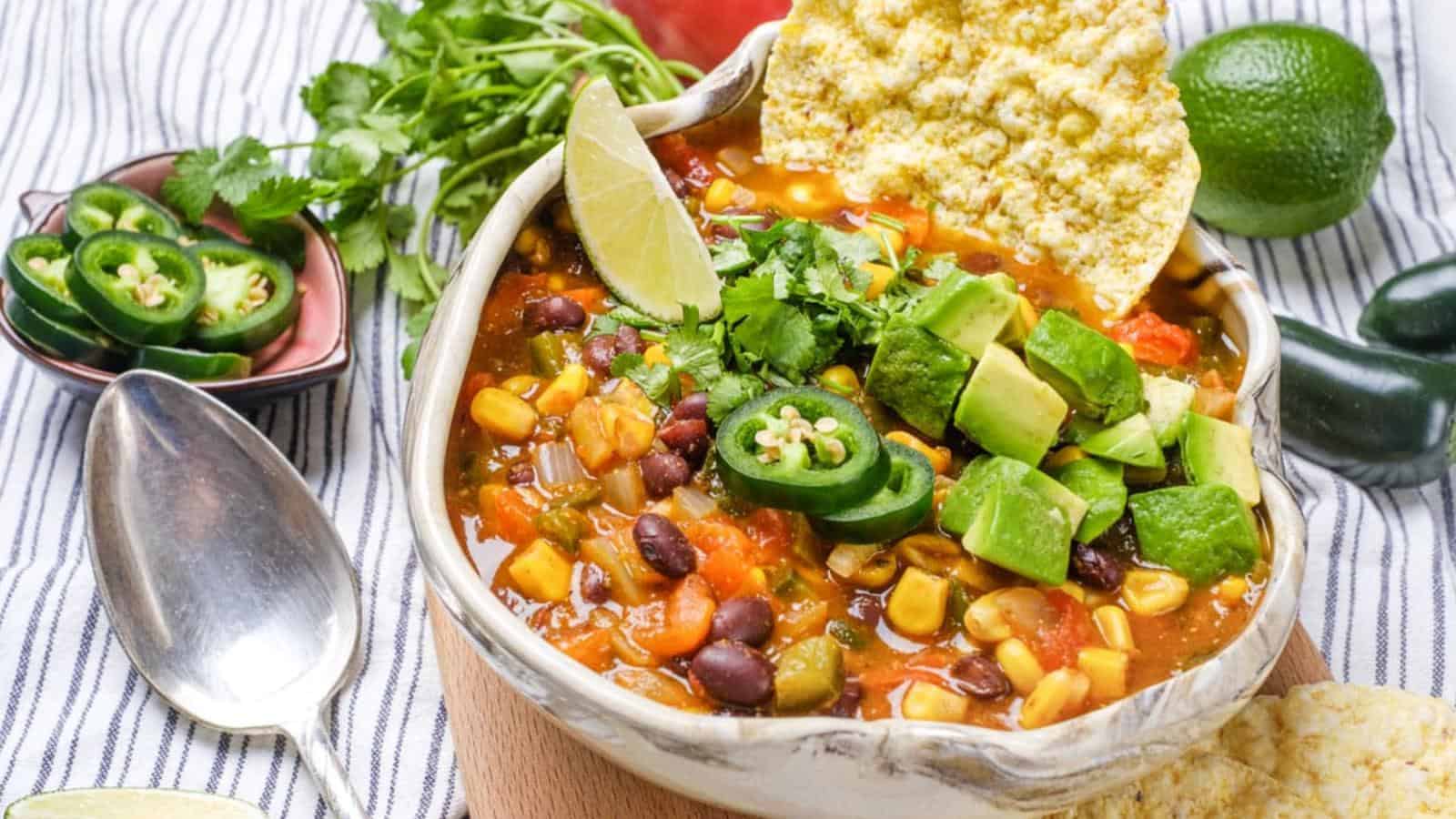 A bowl of vegetable chili topped with avocado, cilantro, jalapeño slices, a lime wedge, and a tortilla chip, with a spoon and fresh ingredients on the side.