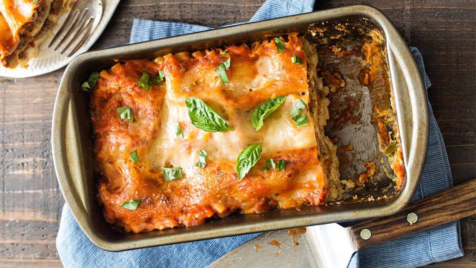 A rectangular baking dish with partially sliced lasagna topped with tomato sauce, melted cheese, and basil leaves on a wooden table.