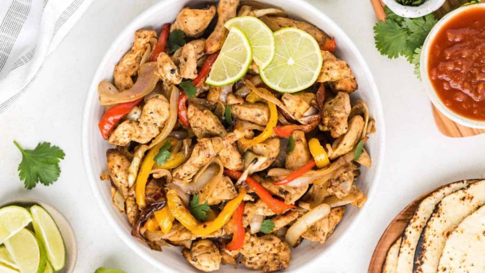 A white bowl filled with cooked chicken strips, red and yellow bell peppers, and onions, garnished with lime slices. Tortillas, salsa, and lime wedges are on the side.