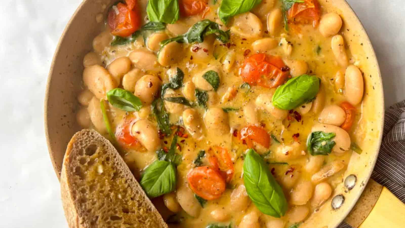 A skillet filled with creamy white beans, cherry tomatoes, spinach, fresh basil, and chili flakes, served with a slice of toasted bread.