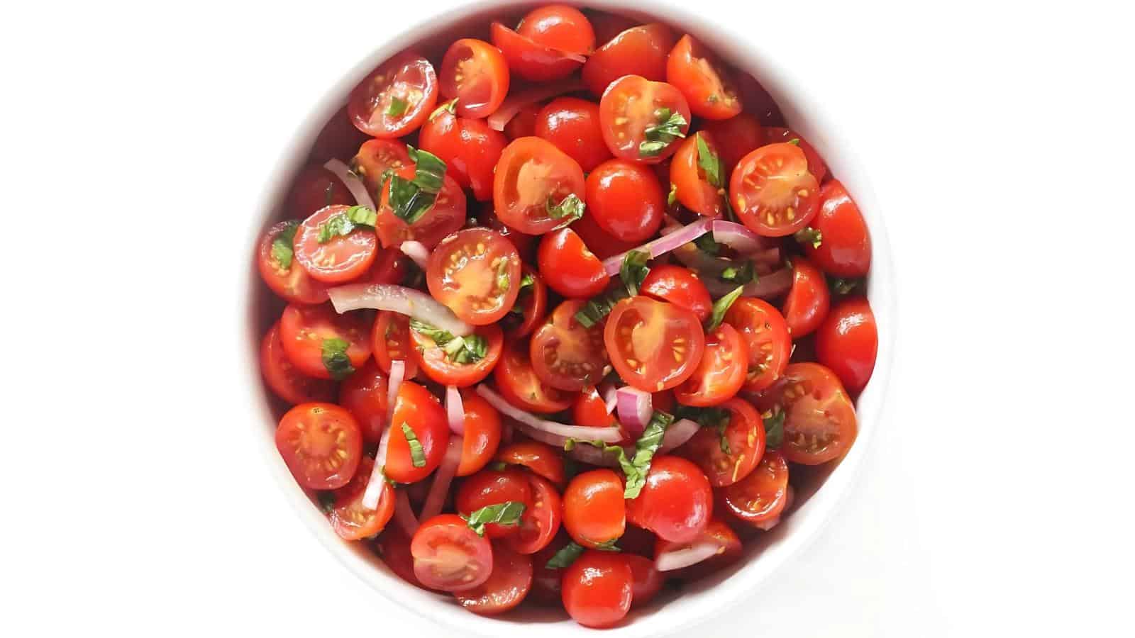 A white bowl filled with halved cherry tomatoes, sliced red onions, and chopped fresh herbs, viewed from above on a white background.