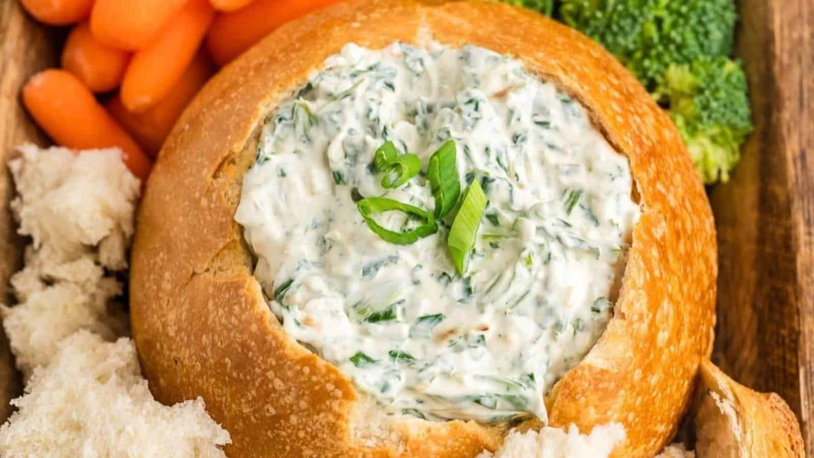 A bread bowl filled with spinach dip, garnished with sliced green onions, surrounded by baby carrots, broccoli, and pieces of bread for dipping.