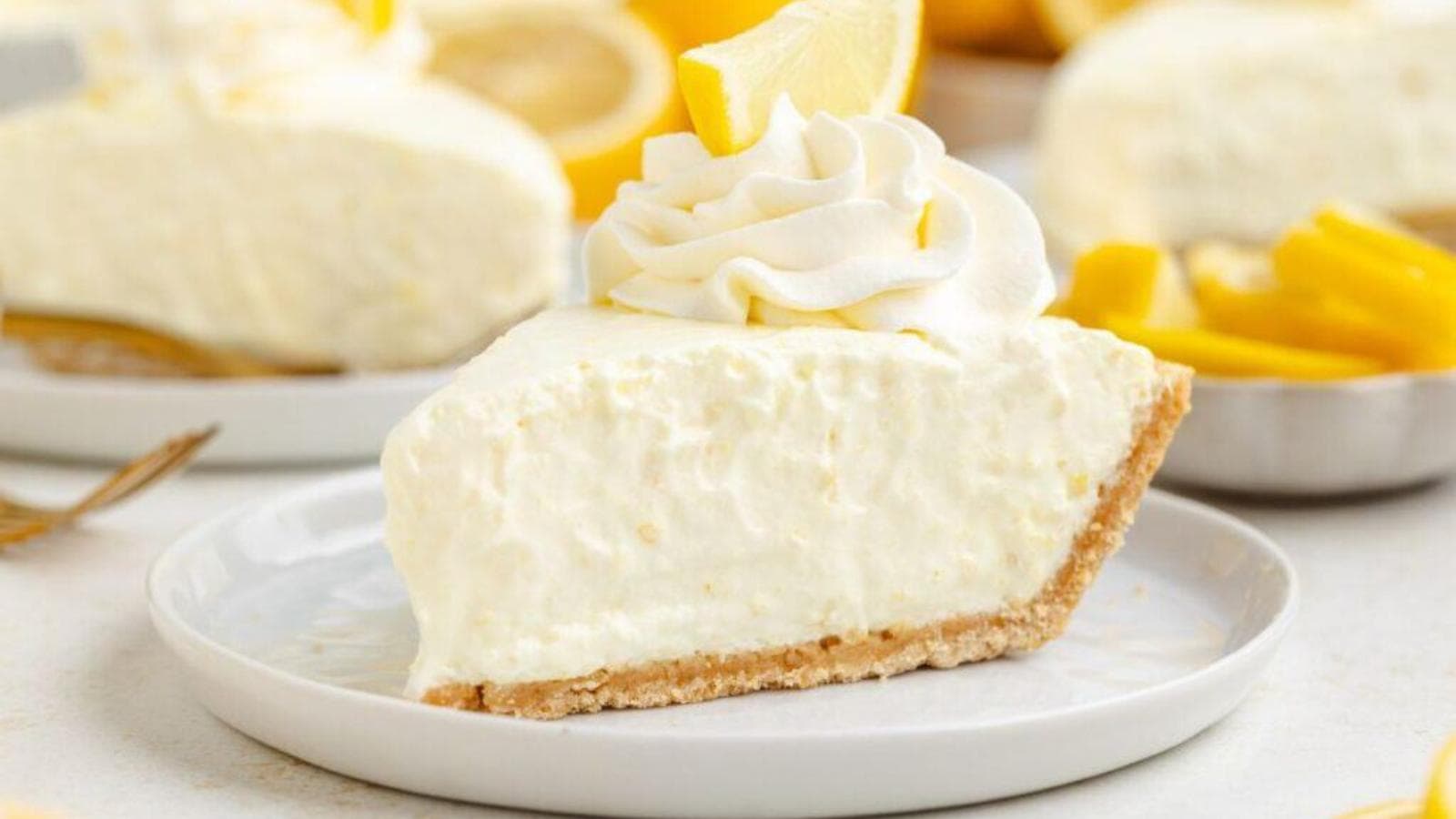 A slice of creamy lemon pie with a graham cracker crust, topped with whipped cream and a lemon wedge, is served on a white plate.