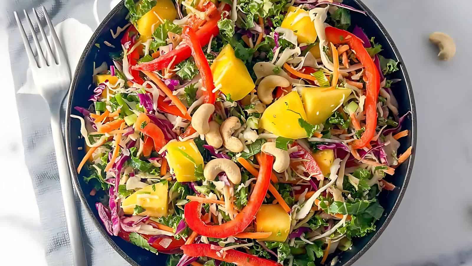 A bowl of colorful salad with chopped kale, red cabbage, carrot, red bell pepper, mango chunks, cashews, and cilantro. A fork and knife are placed beside the bowl.