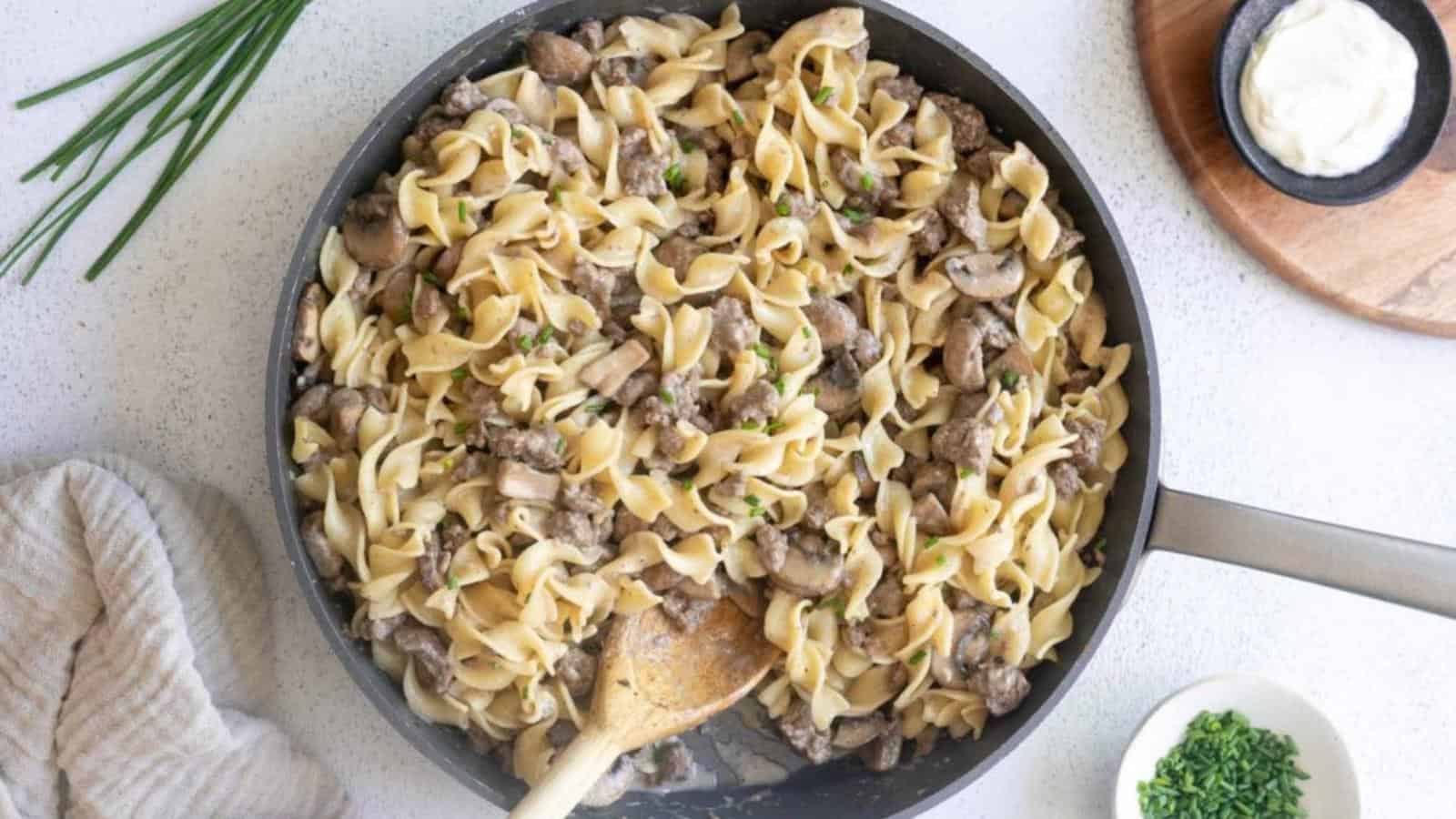 A skillet filled with beef stroganoff made with egg noodles, mushrooms, and beef, garnished with chopped herbs; a wooden spoon rests inside the pan.
