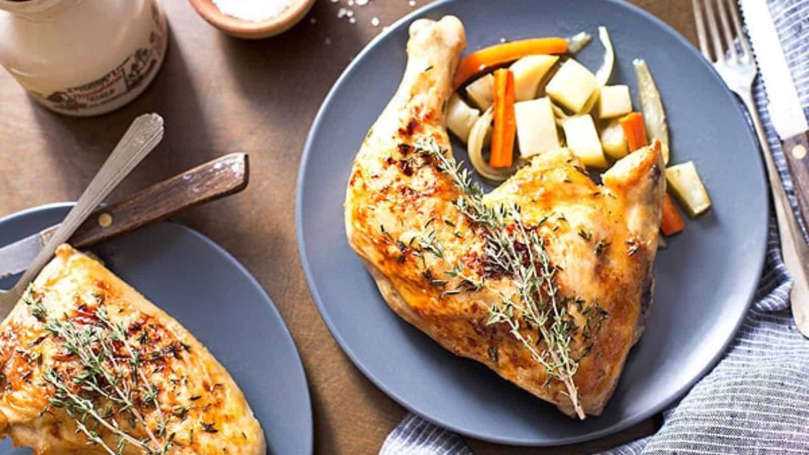 Roast chicken leg served on a gray plate with roasted root vegetables and garnished with fresh thyme. Another plate with chicken and a knife are also visible.